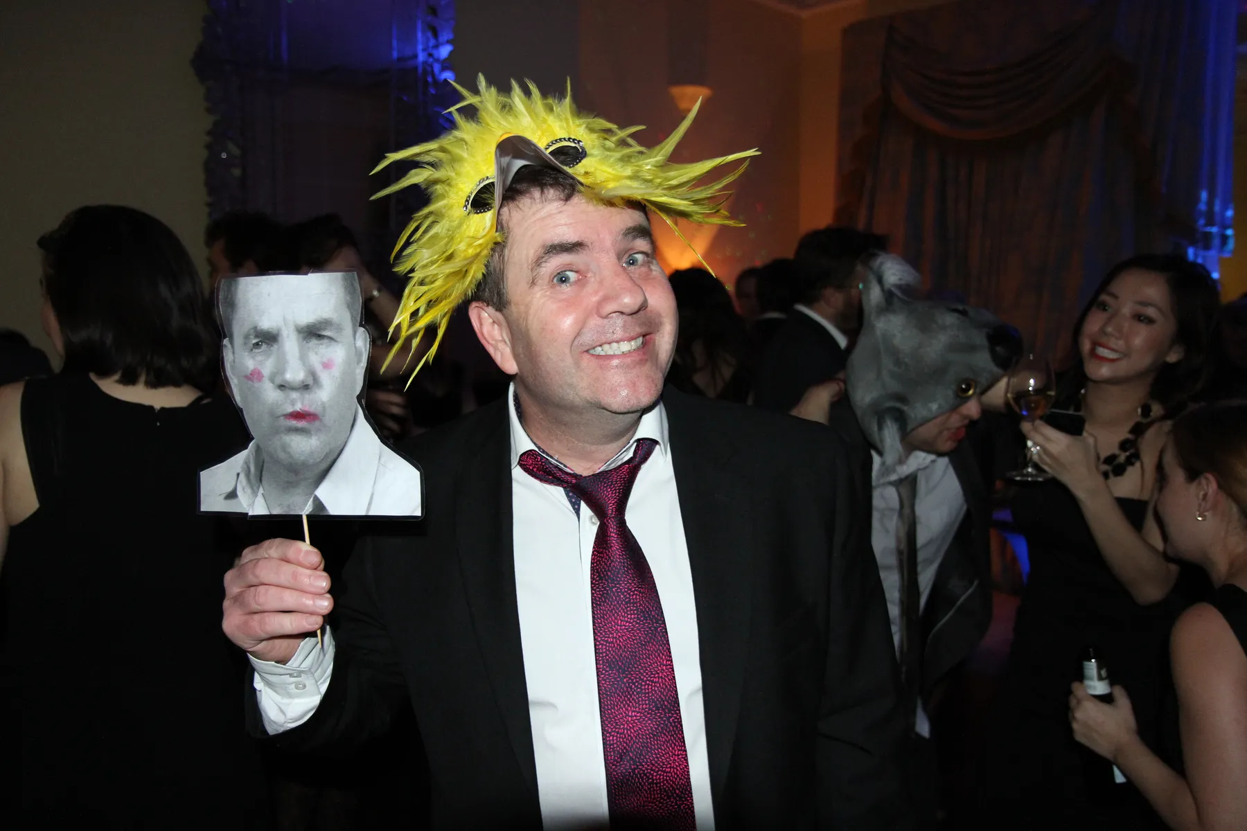 A man in a suit and red tie wears a yellow spiky wig and animal-nose glasses, smiling at a party. He holds a stick with a black-and-white photo of a serious face. People in fancy dress and masks gather as the party DJ plays music in the background.