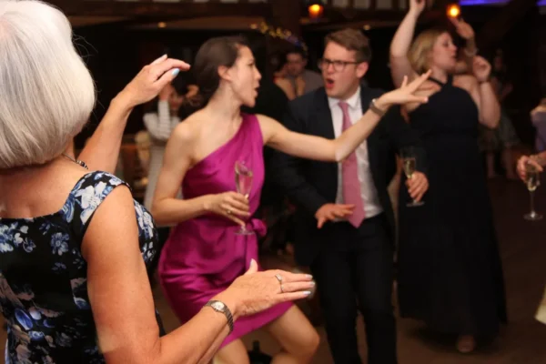 A group of people dressed formally dance enthusiastically at an indoor event with a party DJ. A woman in a purple dress gestures whilst holding a glass, and others around her clap, cheer, and dance on a wooden floor.