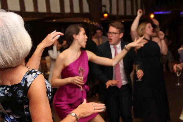 A group of people dressed formally dance enthusiastically at an indoor event with a party DJ. A woman in a purple dress gestures whilst holding a glass, and others around her clap, cheer, and dance on a wooden floor.