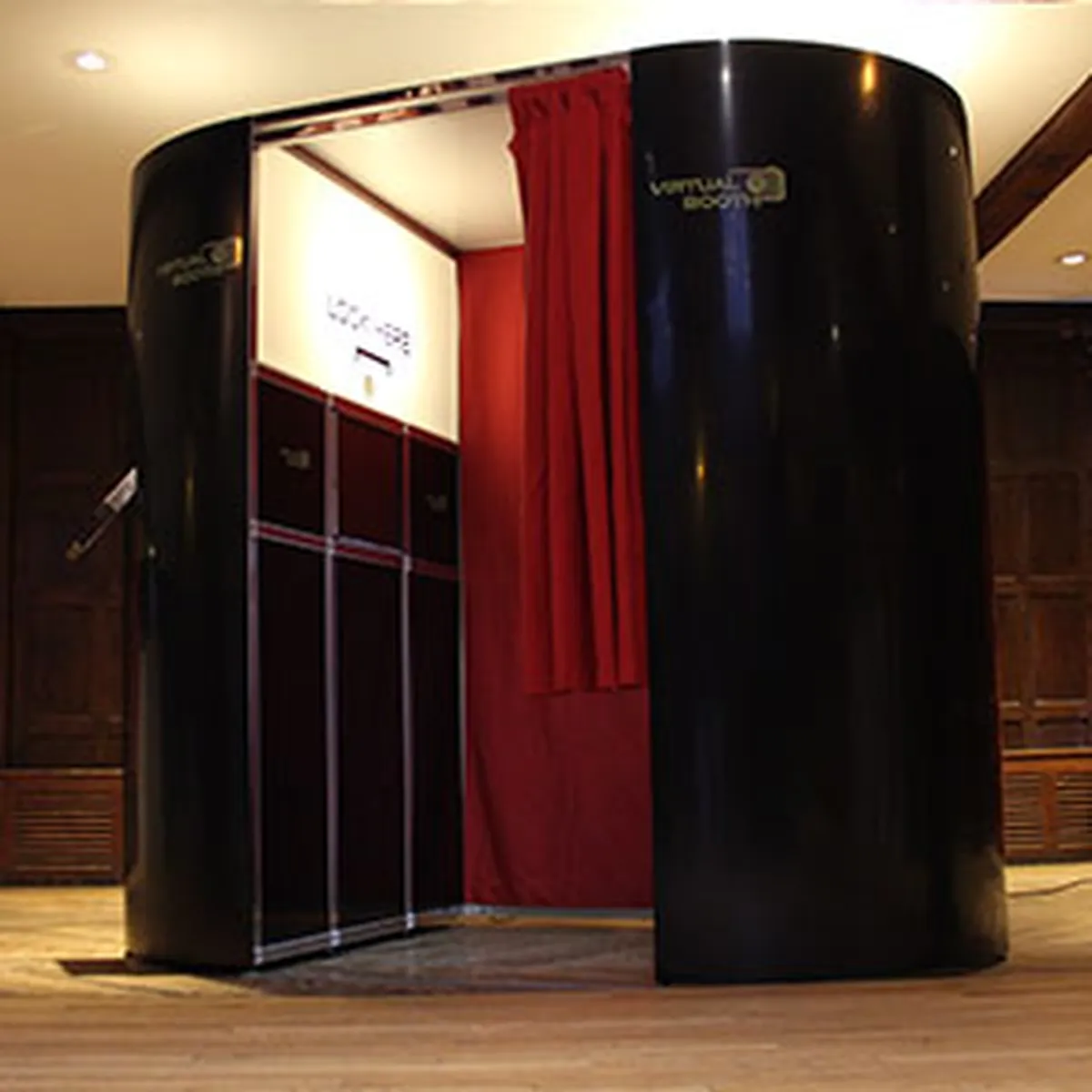 A curved black photo booth with red curtains stands open on a wooden floor in a room with dark wood panelling. Soft lighting highlights the inviting photo booth, with a sign above the entrance.