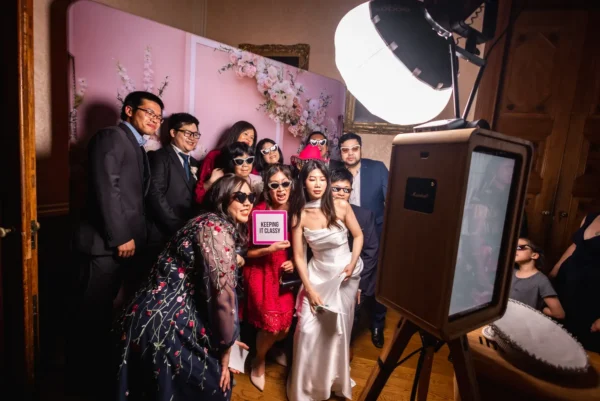 A group of people, including a bride in a white dress, pose together in a photo booth with playful sunglasses and a KEEPING IT CLASSY sign. There’s a pink flower backdrop and a professional photo booth lighting set-up.