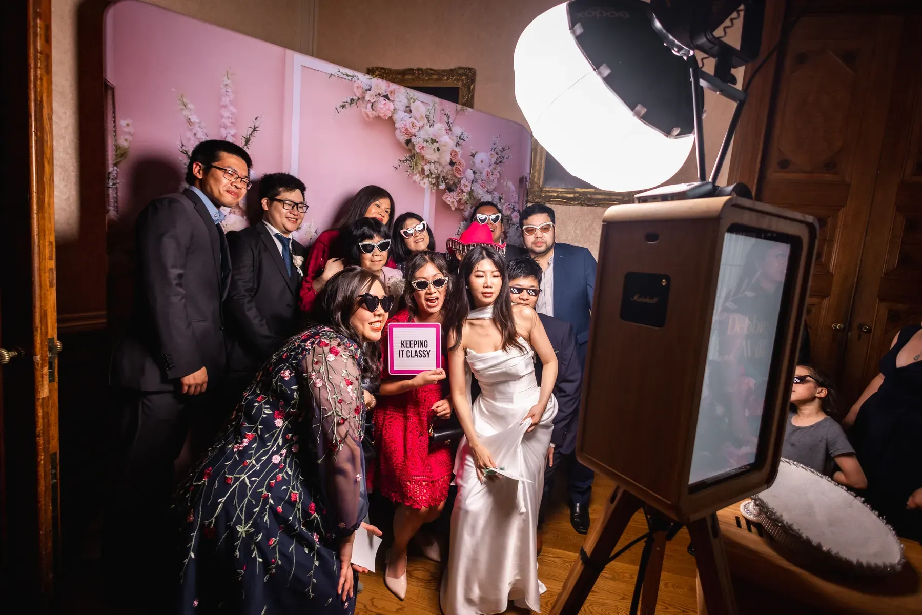 A group of people, including a bride in a white dress, pose together in a photo booth with playful sunglasses and a KEEPING IT CLASSY sign. There’s a pink flower backdrop and a professional photo booth lighting set-up.