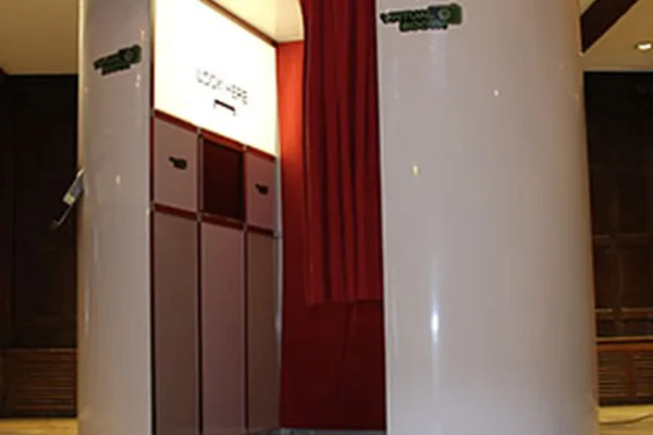 A white, vintage-style photo booth with red curtains stands on a wooden floor in a dimly lit room. The booth’s entrance is partially open, offering a glimpse of the classic photo booth experience behind the curtains.