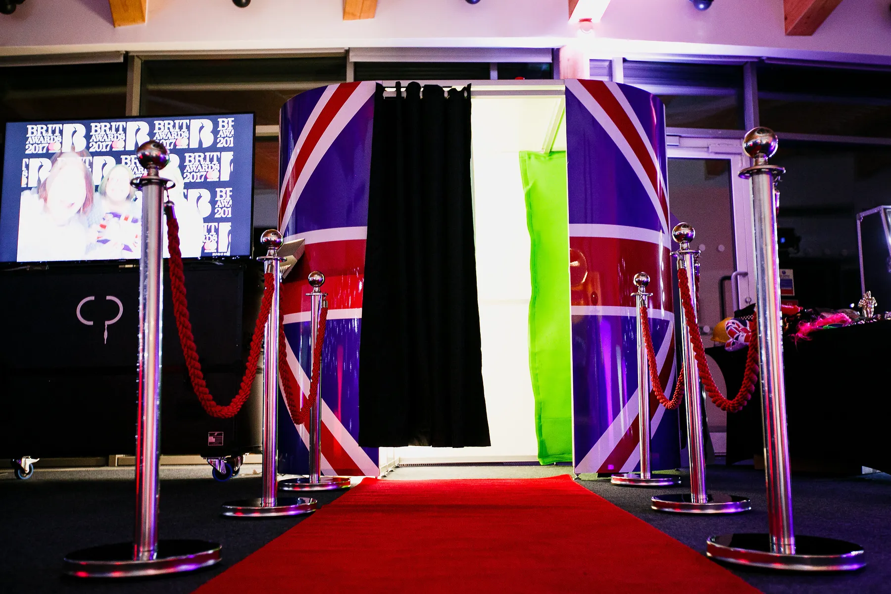 A Union Jack photo booth stands at the end of a red carpet, flanked by silver posts and red ropes, creating a vibrant and inviting indoor photo booth experience.