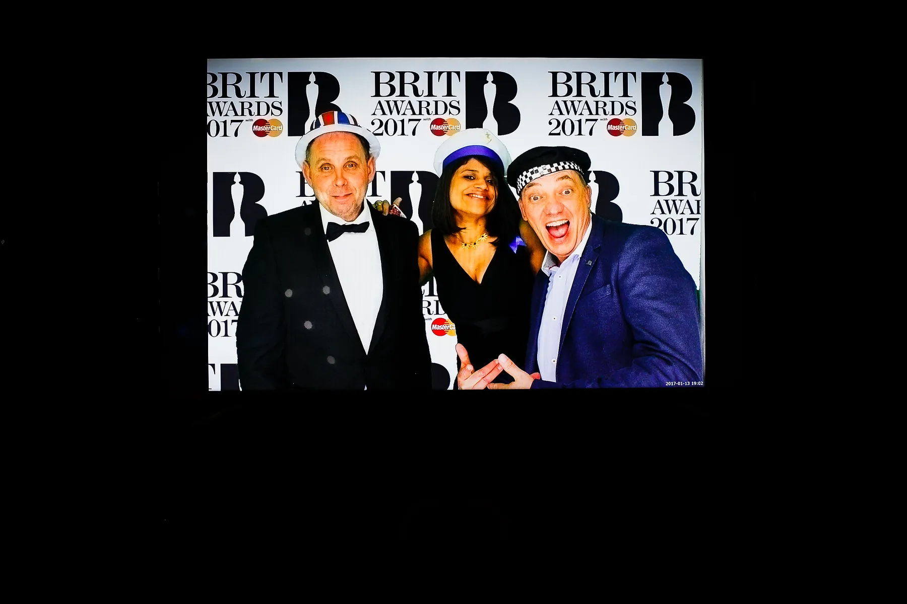 Three people pose and smile as if in a photo booth in front of a Brit Awards 2017 backdrop; two men wear hats and suits, while the woman in the centre wears a black dress and blue hat.