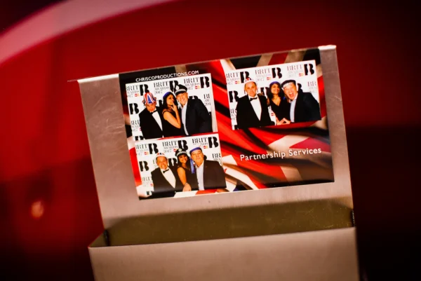 A metallic holder displays a business card featuring three photo booth snapshots of people posing at an event, with a backdrop and the words “Partnership Services” printed on the card. The background is red and blurred.