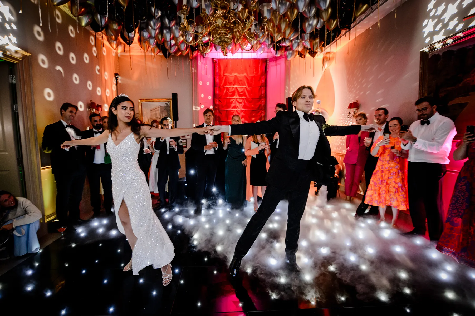A bride and groom perform a ballroom dance in the glamorous wedding venue, Cliveden House, surrounded by guests in formal attire. The dance floor glows with white lights, and balloons and disco dots decorate the ceiling and walls.