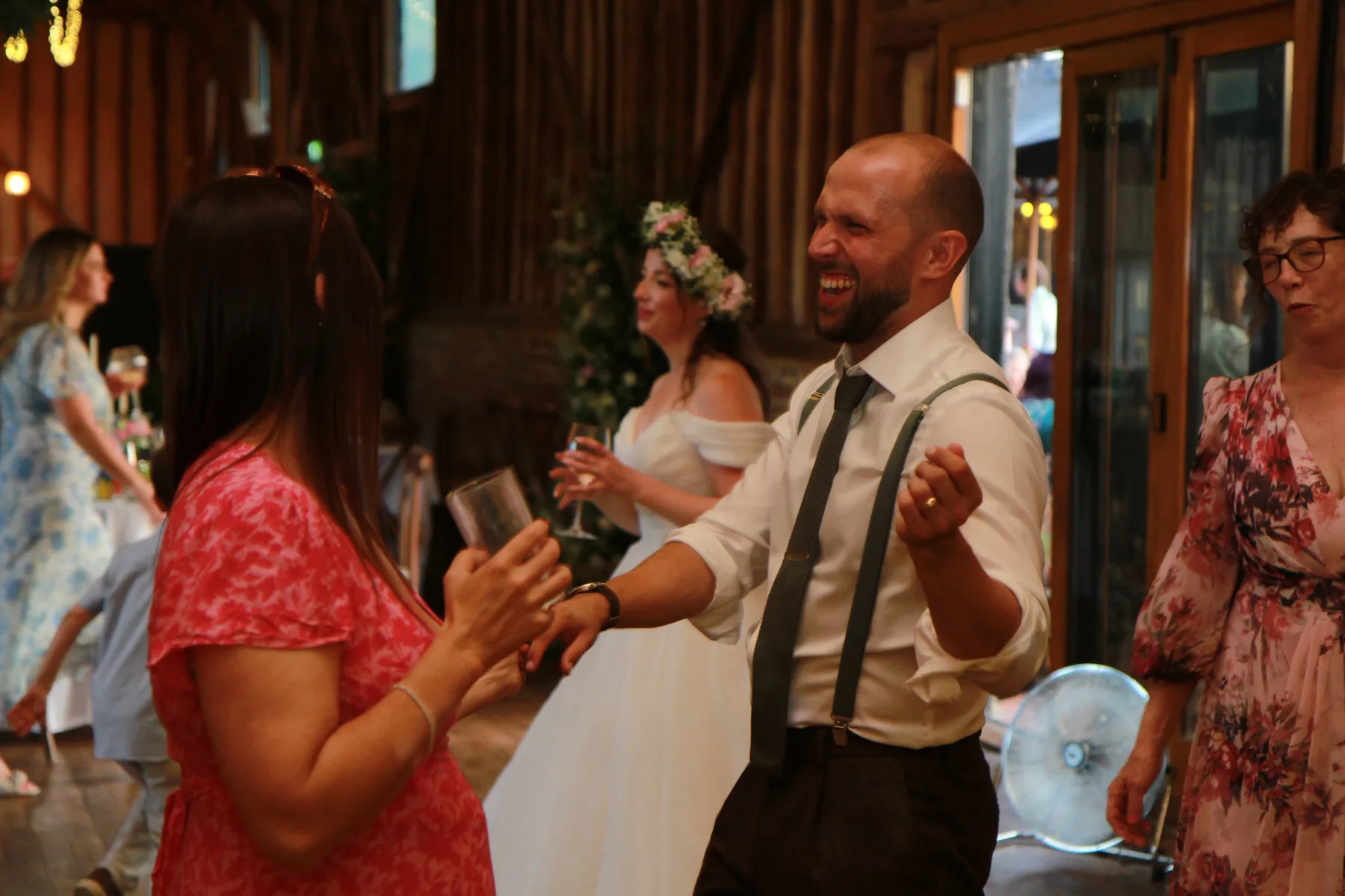 A joyful man in a white shirt and braces dances and laughs with a woman in a pink dress at a rustic indoor wedding, while the wedding DJ Berkshire keeps the energy high and a bride in a white gown and flower crown smiles in the background.