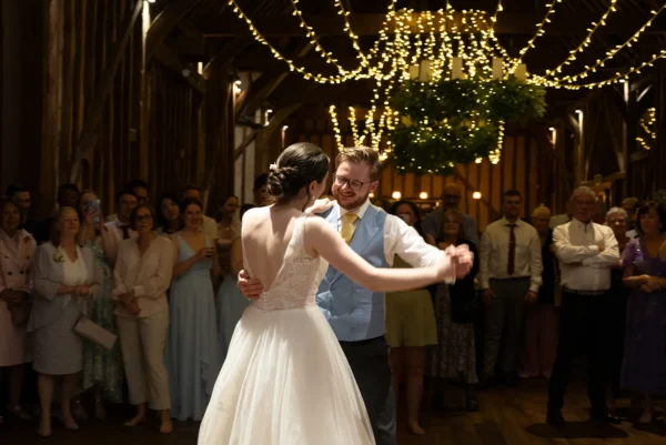A bride and groom share their first dance in a rustic barn decorated with fairy lights, surrounded by smiling guests watching and celebrating their wedding.