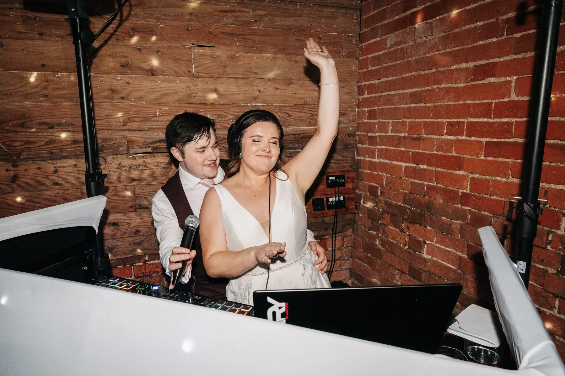 A woman in a white dress wearing headphones joyfully raises her arm while DJing, with a smiling man standing closely behind her. They are in a cosy room with brick and wooden walls.