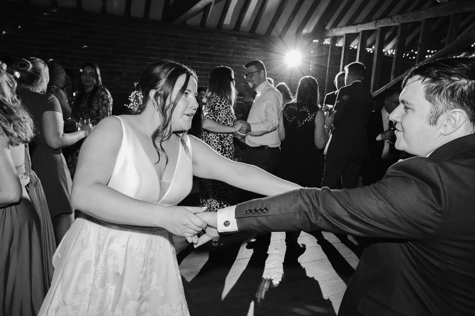 A bride and groom joyfully dance together at their wedding reception at Stanlake Park, Berkshire, surrounded by guests in a barn venue with fairy lights and wooden beams, captured in black and white.