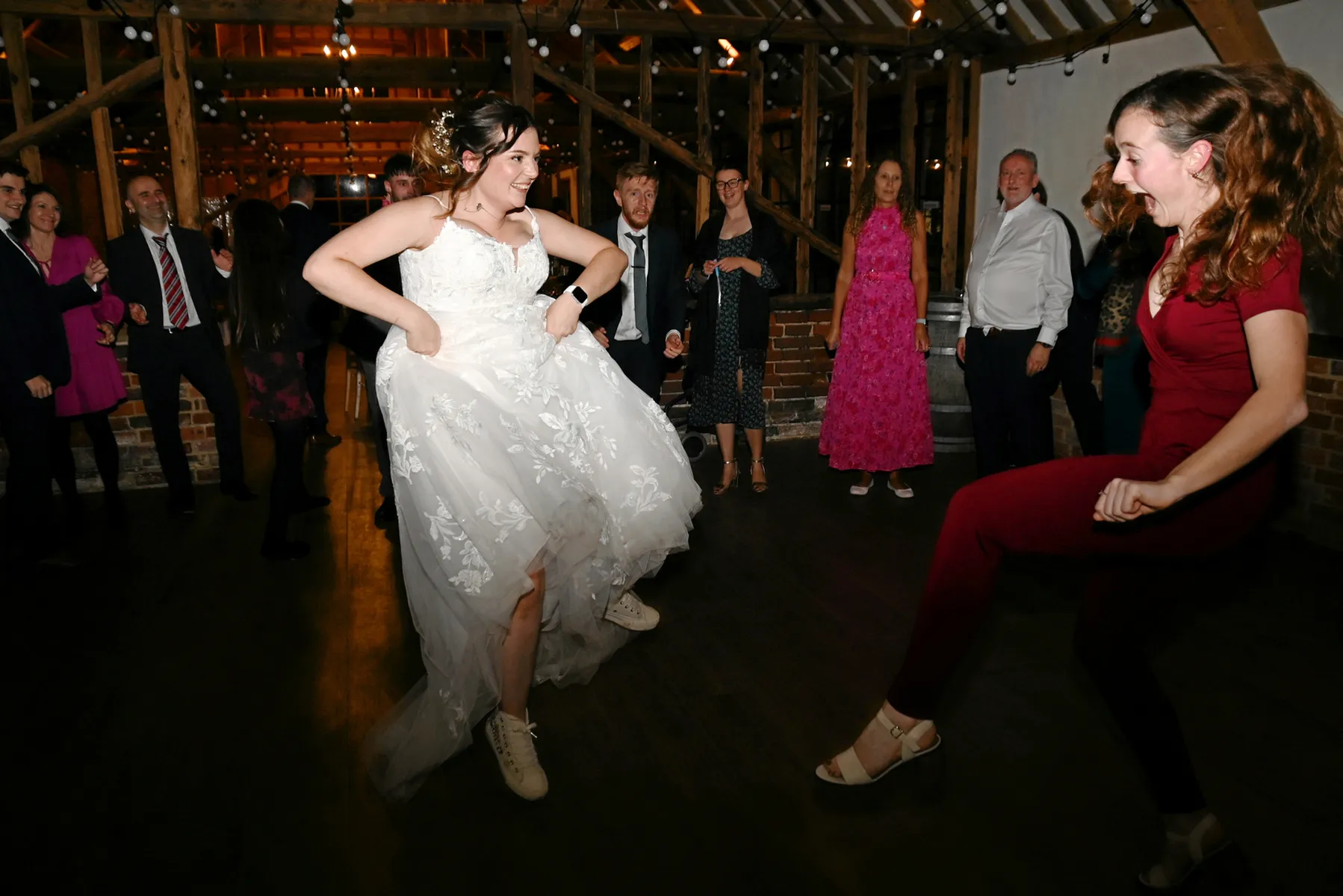 A bride in a white dress and trainers dances enthusiastically with a woman in a red jumpsuit, surrounded by smiling and clapping guests in a warmly lit rustic venue.