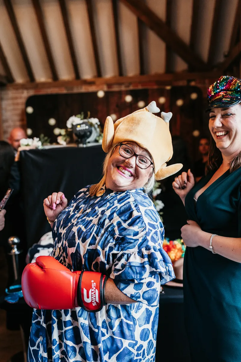 A smiling woman wearing a turkey hat, patterned dress, and red boxing gloves poses playfully at a party, whilst another woman beside her laughs and wears a colourful sequinned mask.