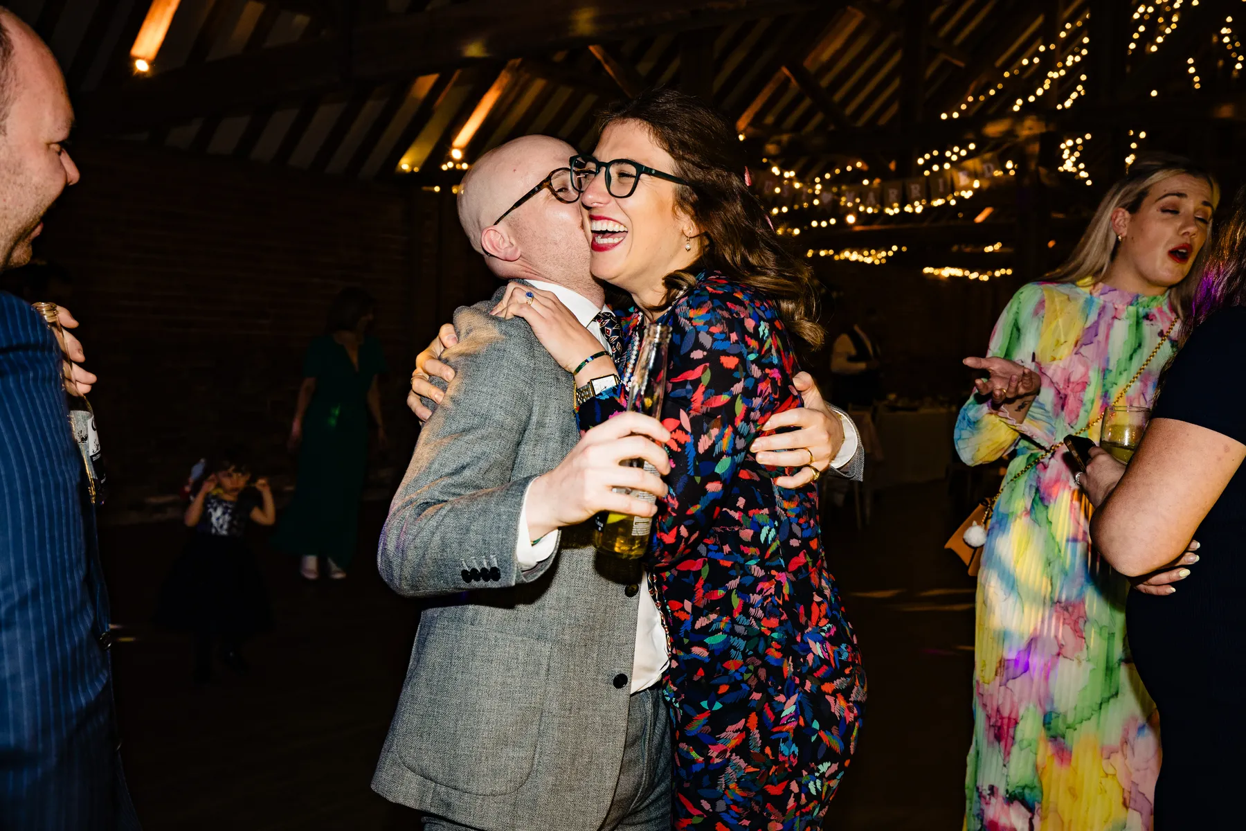 A man in a grey suit and a woman in a colourful outfit hug and laugh on a dance floor at a lively indoor party with fairy lights. Other guests are in the background, some dancing and smiling.