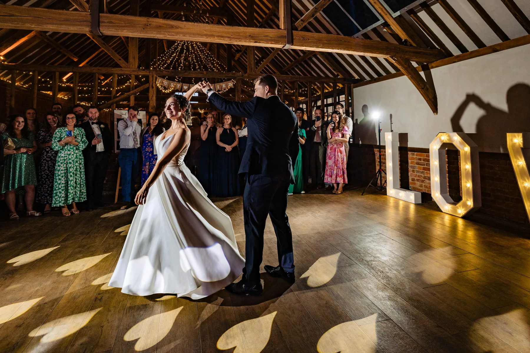 A bride and groom share their first dance in a rustic barn venue with wooden beams, surrounded by guests. The word LOVE is lit up in large letters, casting heart-shaped shadows on the wooden floor.