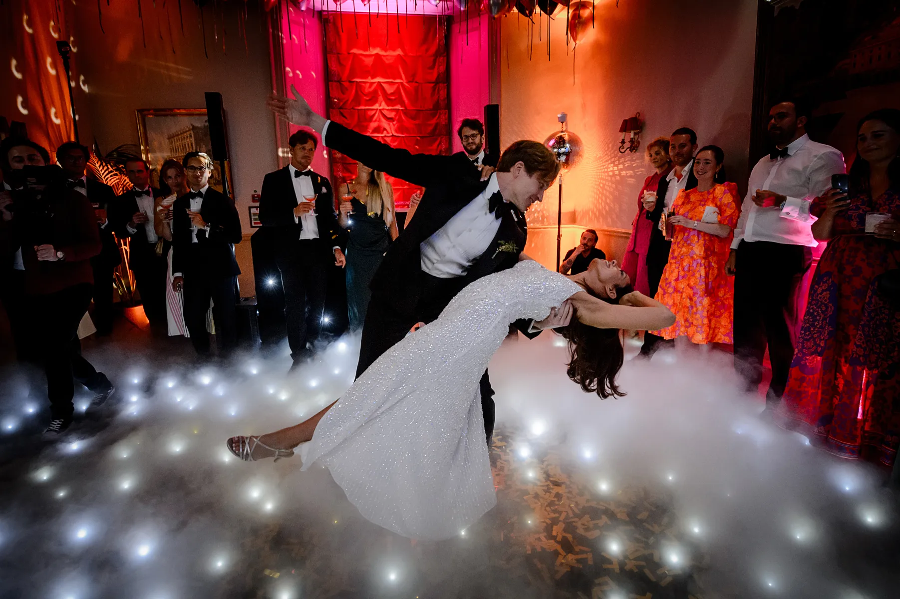 A couple in formal attire performs an elegant dance dip on a misty, illuminated floor, as a wedding DJ Buckinghamshire sets the mood, surrounded by guests in vibrant dresses and dinner jackets at a festive wedding at Cliveden House