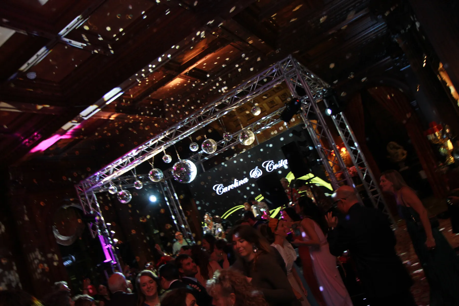 A lively crowd dances under disco balls and colourful lights at a wedding party at luxury wedding venue, Clivden House, Buckinghamshire. The stage is labelled with their names and a wedding DJ keeps the festive atmosphere alive amid sparkling reflections and a decorated ceiling.