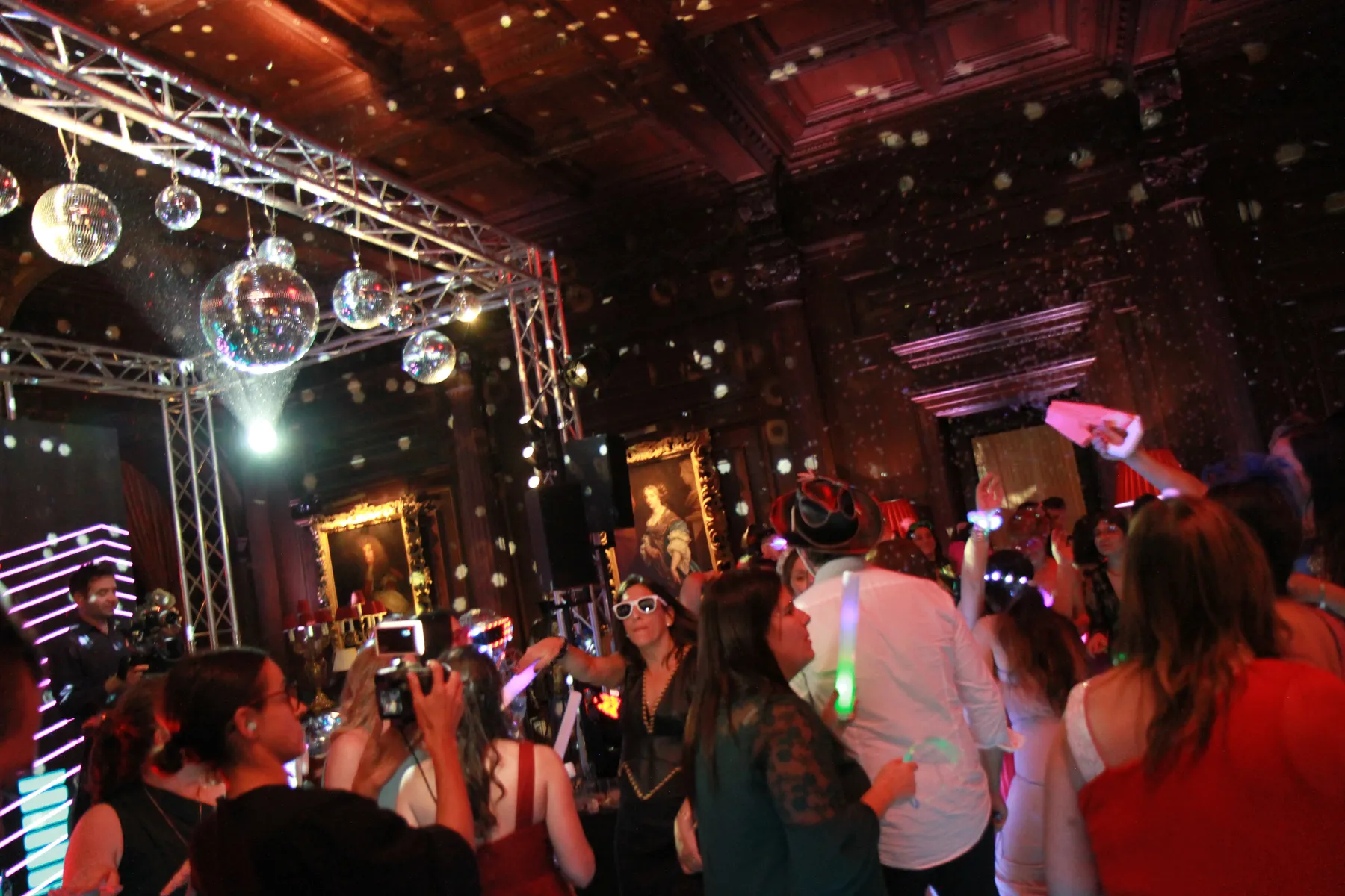 A lively party scene with people dancing under disco balls and colourful lights, brought to life by a wedding DJ Buckinghamshire, in a grand room with dark wood panelling and framed portraits. Guests take photos, wearing hats and glow accessories.
