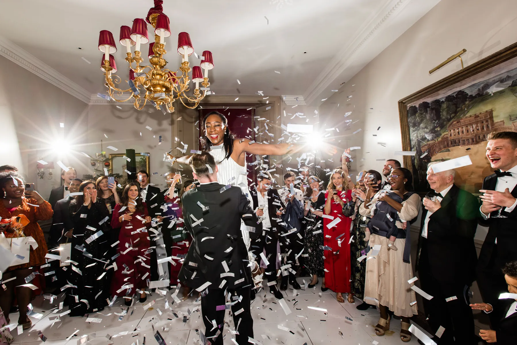 A joyful wedding crowd claps and cheers as confetti falls. A wedding DJ at Cliveden House in Buckinghamshire brings energy to the festivities as the groom lifts his delighted partner, with bright lights and elegant décor enhancing the celebratory atmosphere.