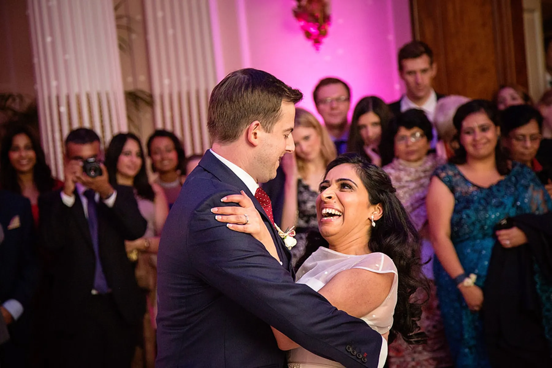 A bride and groom share a joyful first dance at their wedding reception, smiling and gazing at each other, as a wedding DJ Buckinghamshire provides the perfect soundtrack for their celebration, surrounded by cheering and happy guests in formal attire.