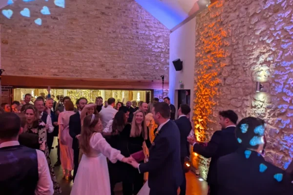 A lively wedding reception in a rustic stone hall with fairy lights and a top wedding DJ Buckinghamshire. Guests, including the bride in white, dance and mingle under colourful lighting and blue heart-shaped light projections.