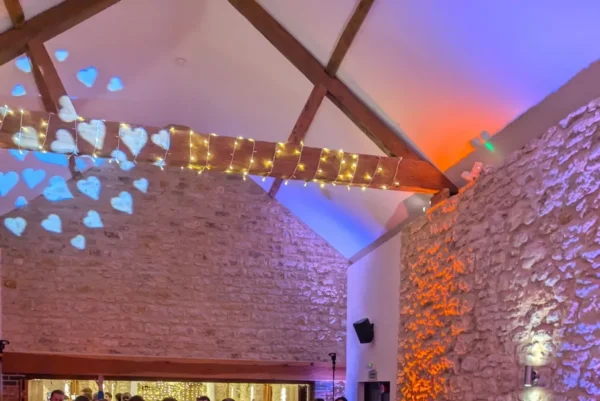 A lively wedding reception in a rustic stone hall with fairy lights and a top wedding DJ Buckinghamshire. Guests, including the bride in white, dance and mingle under colourful lighting and blue heart-shaped light projections.