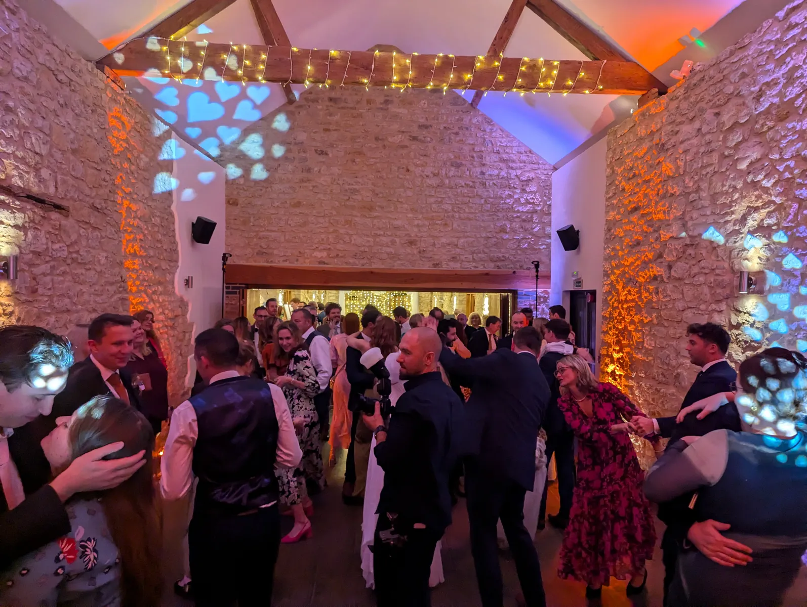 A large group of people, dressed in formal attire, dance and socialise in a warmly lit stone hall decorated with fairy lights and colourful projections—perfectly set for a celebration with a talented wedding DJ at Huntsmill Farm in Buckinghamshire.