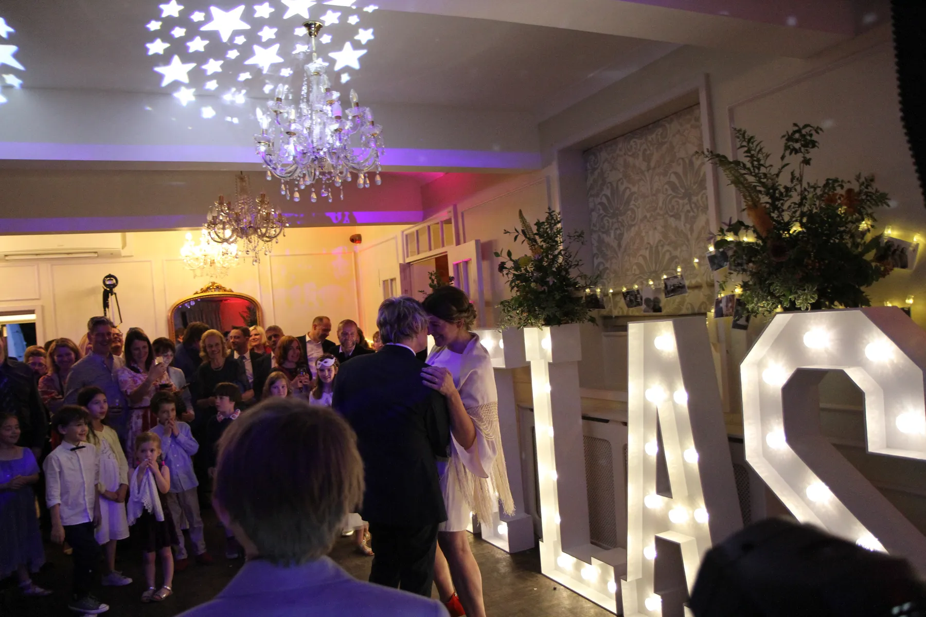 A couple dances in a warmly-lit room decorated at Kings Chapel in Old Amersham, with large illuminated letters, whilst guests, including children, watch and smile. A chandelier and star-shaped lights add to the festive atmosphere, perfectly set by a talented wedding DJ Buckinghamshire.
