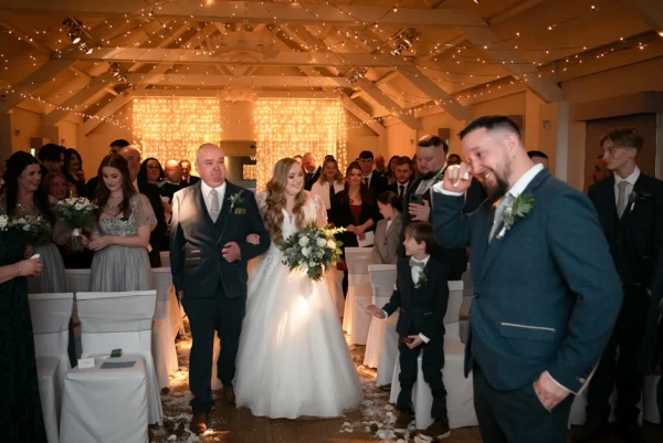 A bride walks down the aisle with her father as guests look on and the groom wipes away a tear; fairy lights and music from a top wedding DJ Buckinghamshire create a warm, festive atmosphere in the venue.