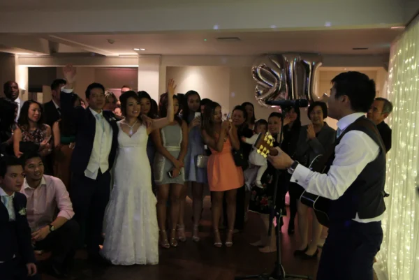 A bride and groom stand together smiling in front of a crowd at their wedding reception whilst a man plays guitar and sings. Guests are watching, some with raised hands, and large 21 balloons are visible—perfect for a wedding DJ Buckinghamshire event.