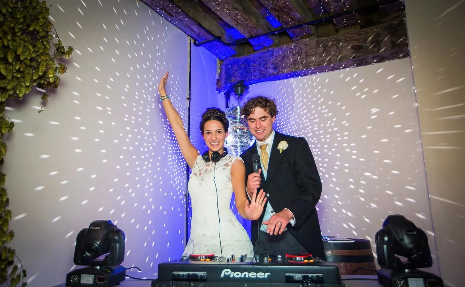 A bride and groom stand behind DJ equipment, smiling. The bride raises one arm and holds headphones, whilst the groom waves. Sparkling lights create a festive atmosphere—perfect for a memorable wedding DJ Essex celebration.