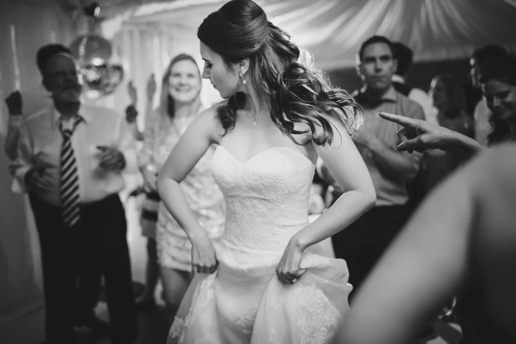 A bride in a strapless wedding dress dances and holds up her gown in a lively, crowded reception as the wedding DJ Essex keeps guests clapping and celebrating round her.