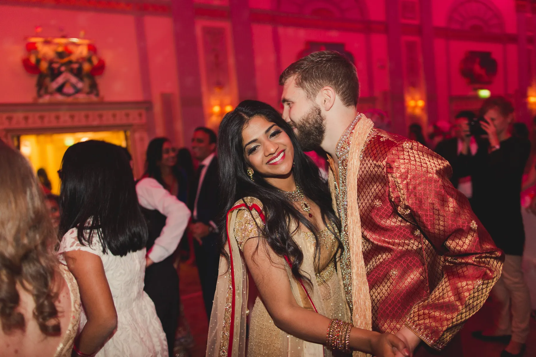 A woman in a gold saree smiles as a man in a red traditional outfit kisses her cheek at a festive indoor event, with guests dancing in the background under warm, pink lighting by a lively wedding DJ Essex.