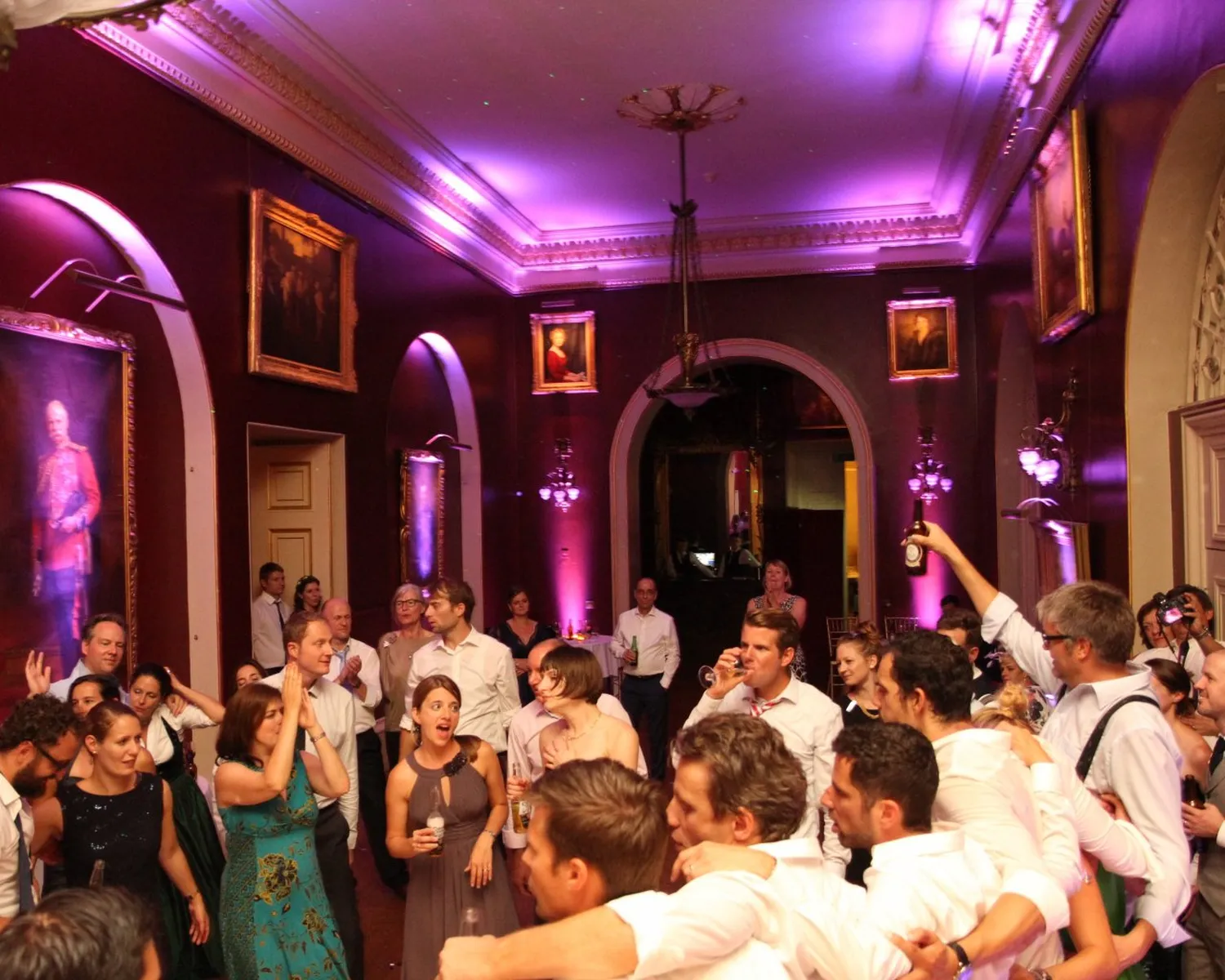 A large group of people in formal attire dance and celebrate to the beats of a wedding DJ Essex in an elegant, purple-lit room with framed portraits on the walls and arched doorways.