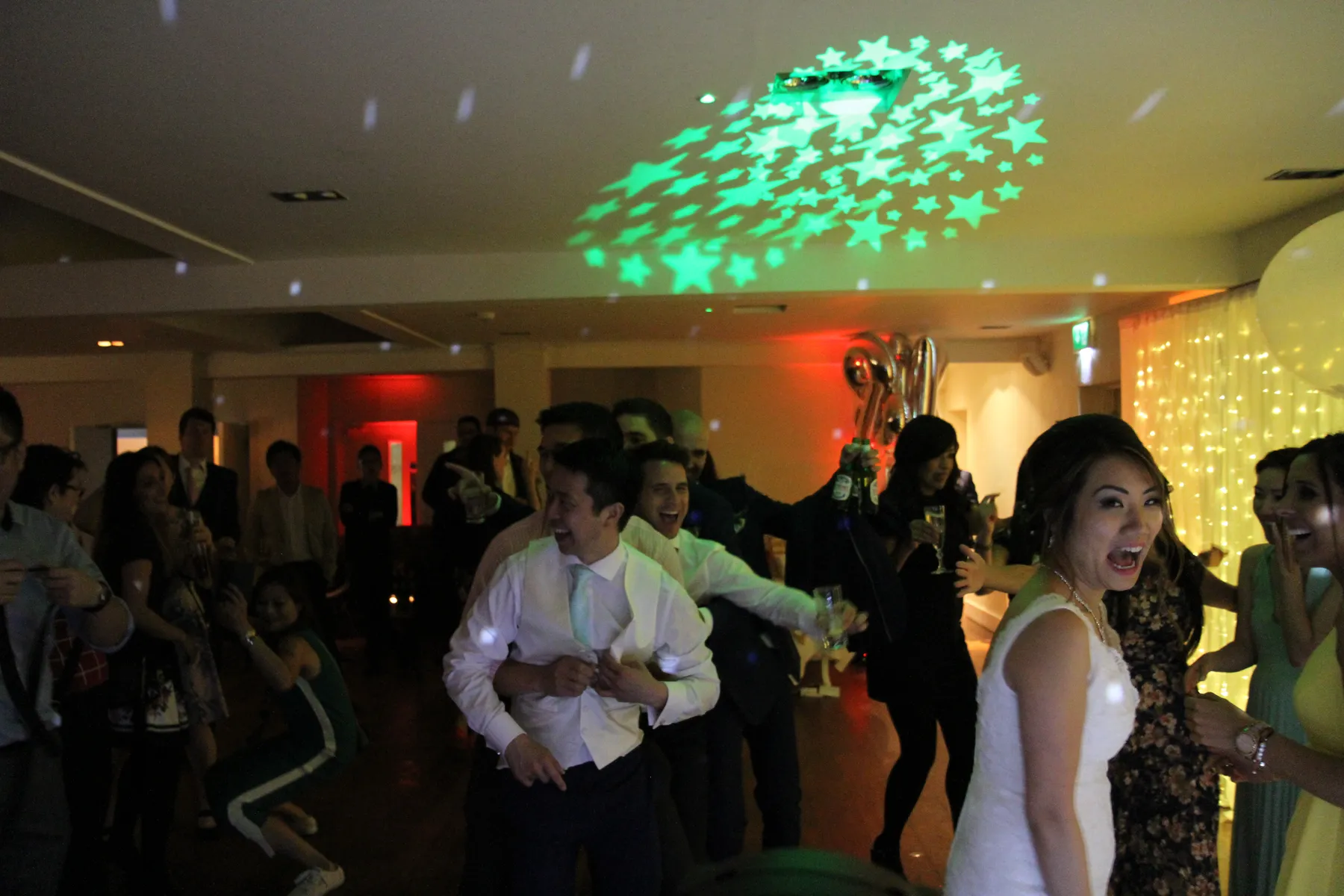 A lively wedding party scene indoors, with guests dancing and laughing under green star-shaped lights from a wedding DJ Essex. The bride, in a white dress, smiles excitedly in the foreground.