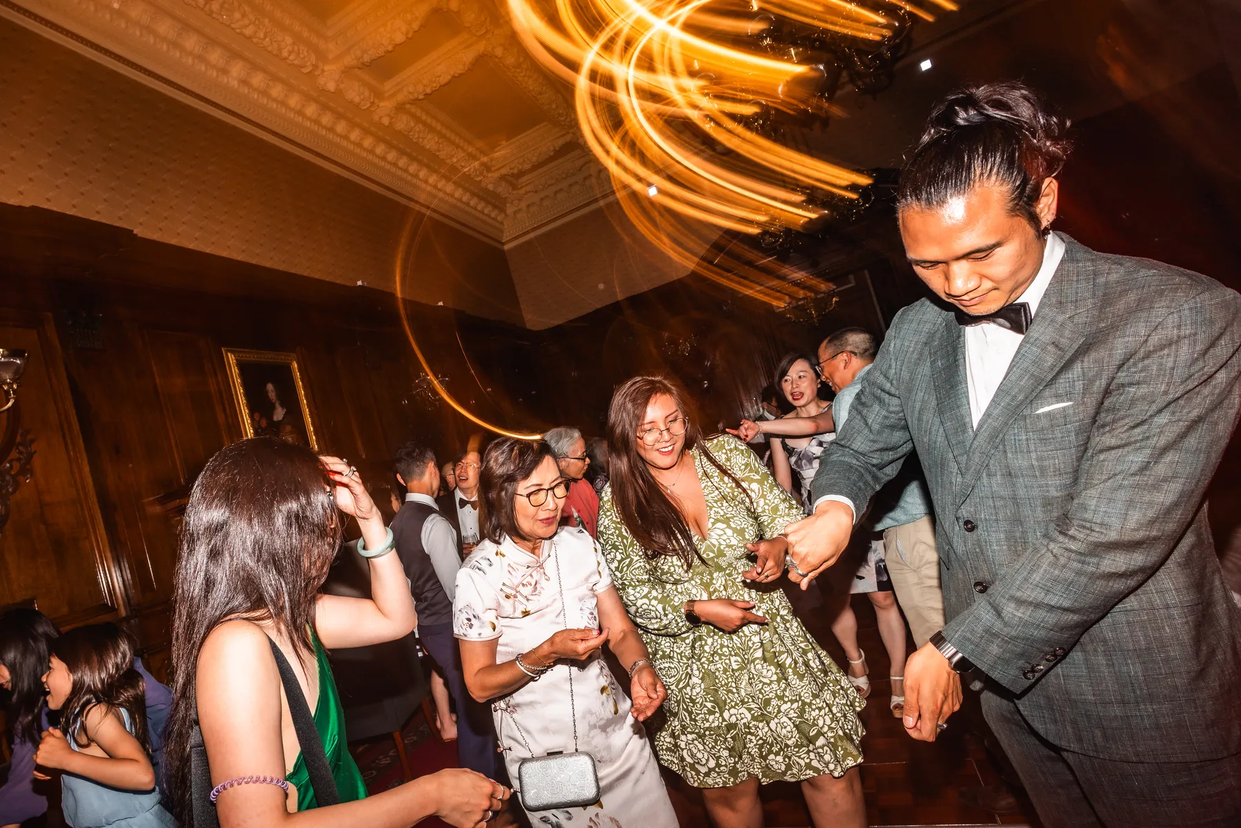 A group of people in formal and semi-formal attire smile and laugh together in a warmly lit, ornate room with wooden walls as swirling light trails from a wedding DJ Essex create a festive atmosphere.