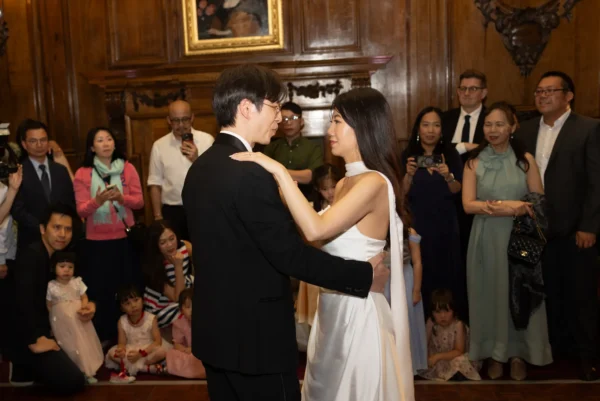 A bride and groom share a dance in a wood-panelled room, surrounded by seated and standing guests, including children and adults who are watching and taking photos.