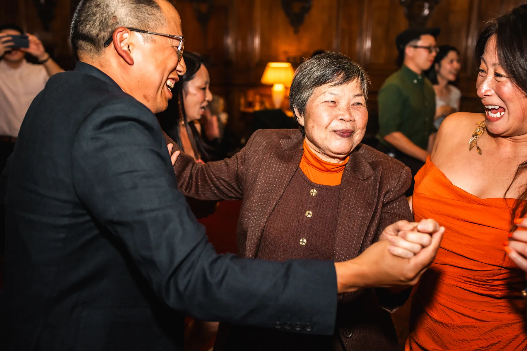 A group of people, including an elderly woman in a brown jacket and others in vibrant outfits, happily dance and hold hands at a lively indoor event. Warm lighting and smiling faces, thanks to a top wedding DJ Hertfordshire, create a joyful atmosphere.