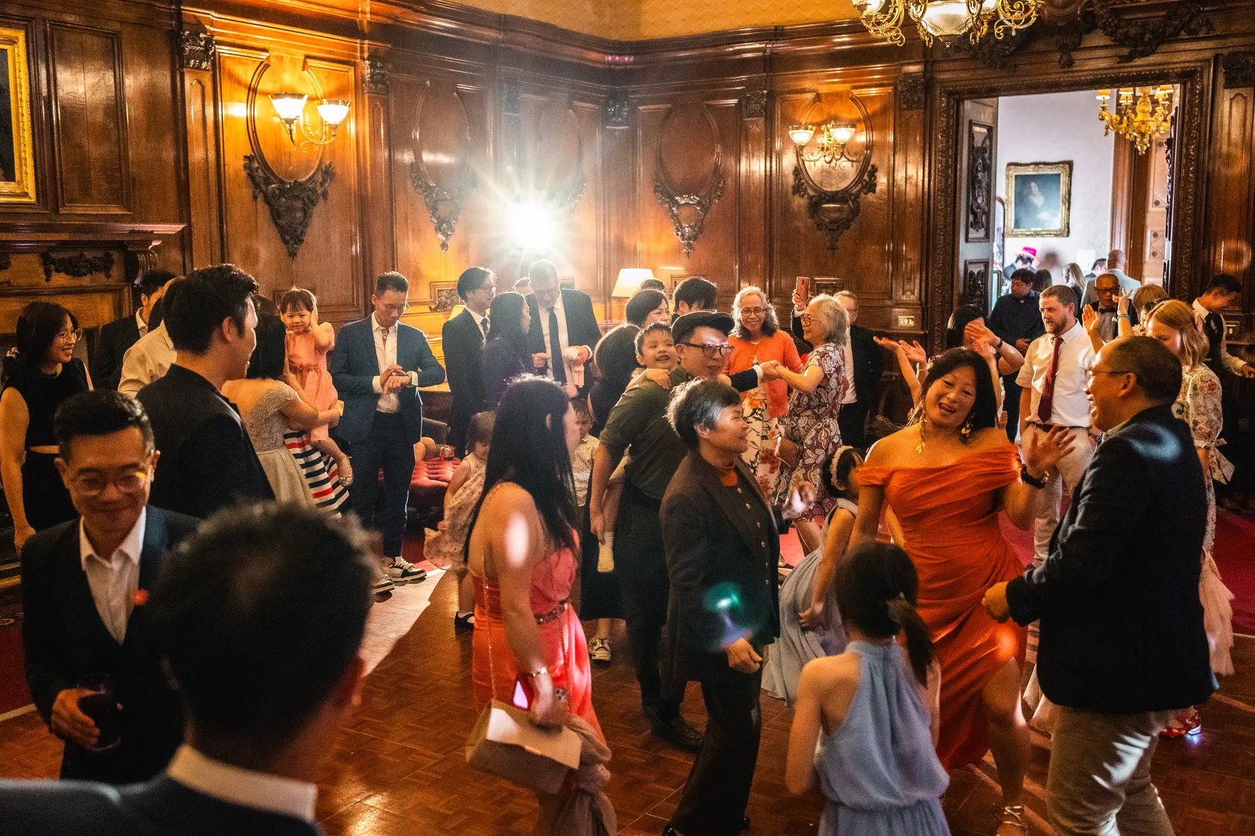 A group of wedding guests in formal attire dance and celebrate in an ornate, wood-panelled room with warm lighting and chandeliers at Ashridge House in Hertfordshire. Some are smiling and laughing, and a bright flash is seen in the background.
