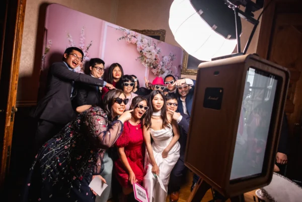 A group of people, including a bride in a white dress, pose happily with fun sunglasses and props for a photo booth at a wedding, smiling and laughing under bright studio lights after dancing to tunes from the top wedding DJ Hertfordshire offers.