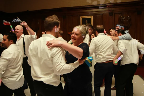 A group of people dressed formally dance and celebrate in a wood-panelled room, enjoying music from a wedding DJ Hertfordshire. A woman in a navy dress dances with a man in a white shirt, while others wave small flags and glow sticks.
