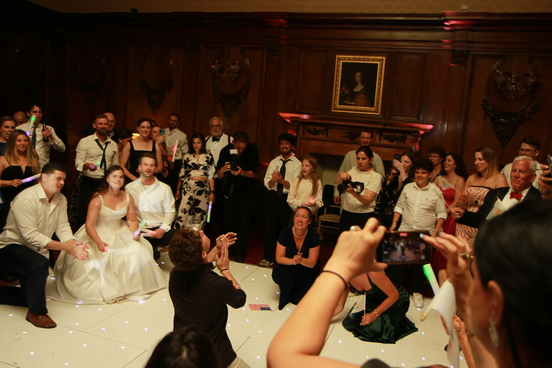 A group of people, including a bride in a white wedding dress, gather and smile on a lit dance floor as the wedding DJ Hertfordshire plays music. Some kneel or crouch, others take photos in a wood-panelled room with a portrait on the wall.