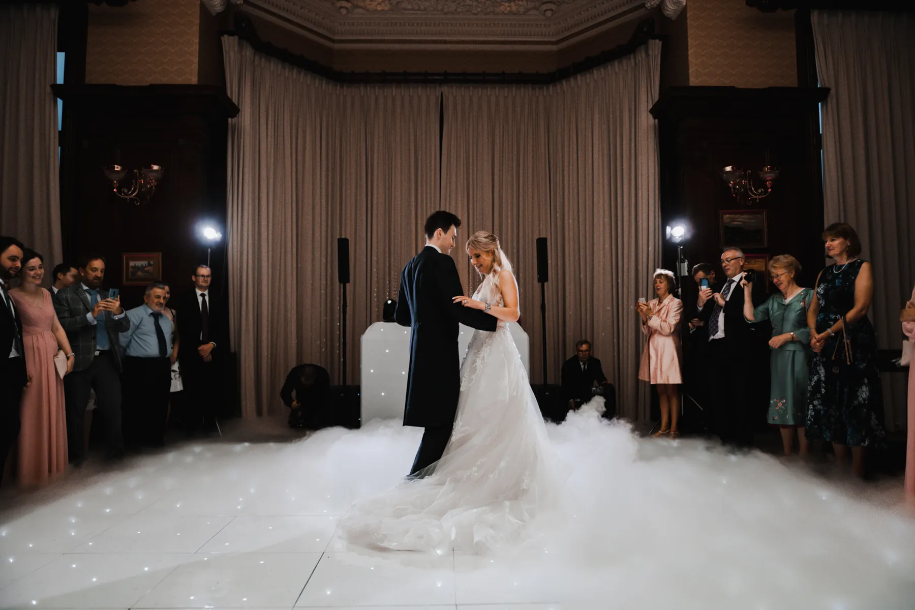 A bride and groom share their first dance on a white, fog-covered floor surrounded by guests in formal attire—an unforgettable moment crafted by a top wedding DJ Hertfordshire in an elegant hall with tall curtains and ornate details.
