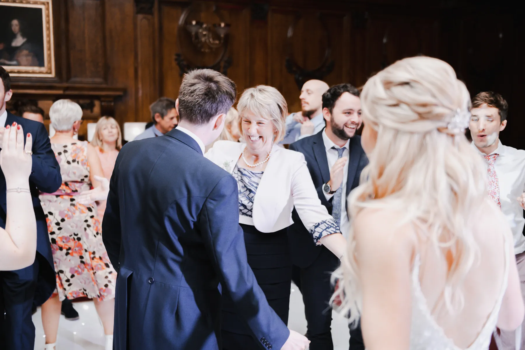A group of people dressed in formal attire dance and smile at a celebration in a wood-panelled room, with a woman in a white jacket in the centre, while a wedding DJ Hertfordshire sets the perfect mood for the joyful event.