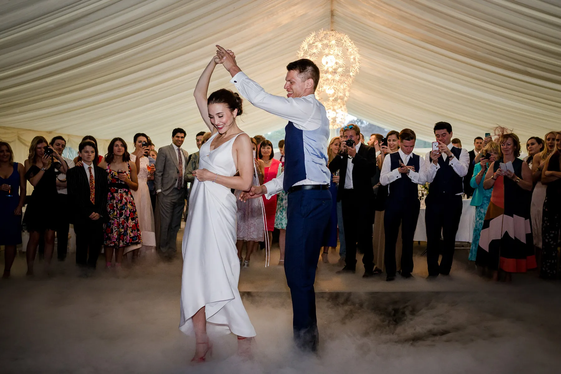 A bride and groom share a first dance on a fog-covered floor in a white marquee, smiling as guests in formal attire watch and take photos. The magical atmosphere was created by their wedding DJ Hertfordshire.