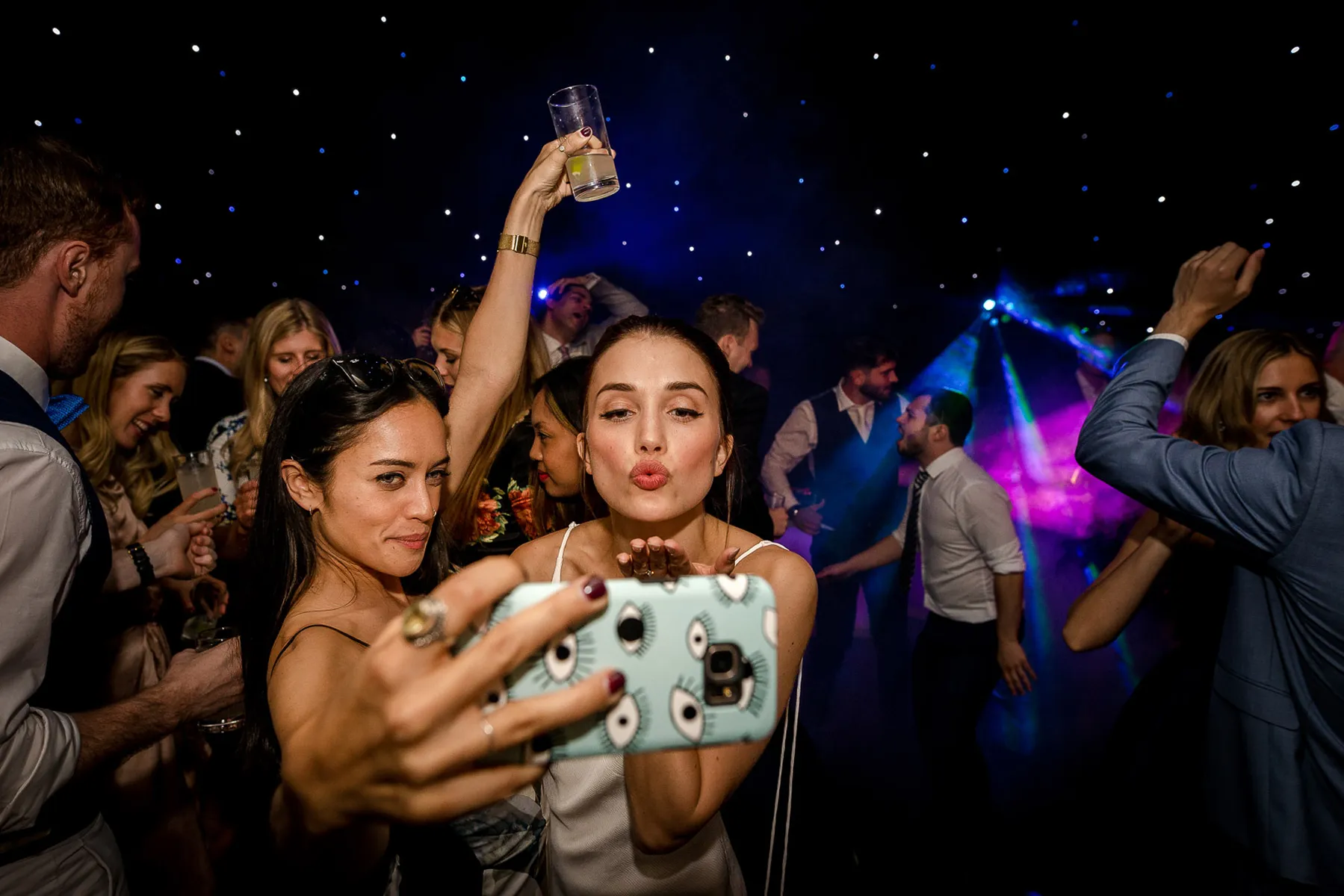 Two women take a selfie at a lively party, one blowing a kiss and the other raising a drink. Colourful lights from the wedding DJ Hertfordshire, people dancing, and a starry backdrop create a festive atmosphere.