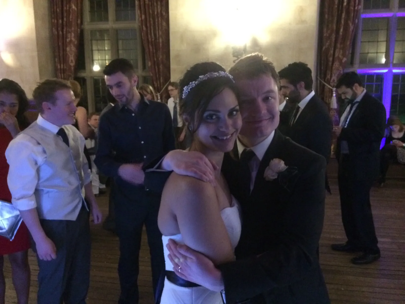 A bride and groom smile whilst embracing on a dance floor, surrounded by wedding guests in formal attire. The decorated hall, with curtains and tall windows, comes alive thanks to a talented wedding DJ Hertfordshire providing the perfect soundtrack.