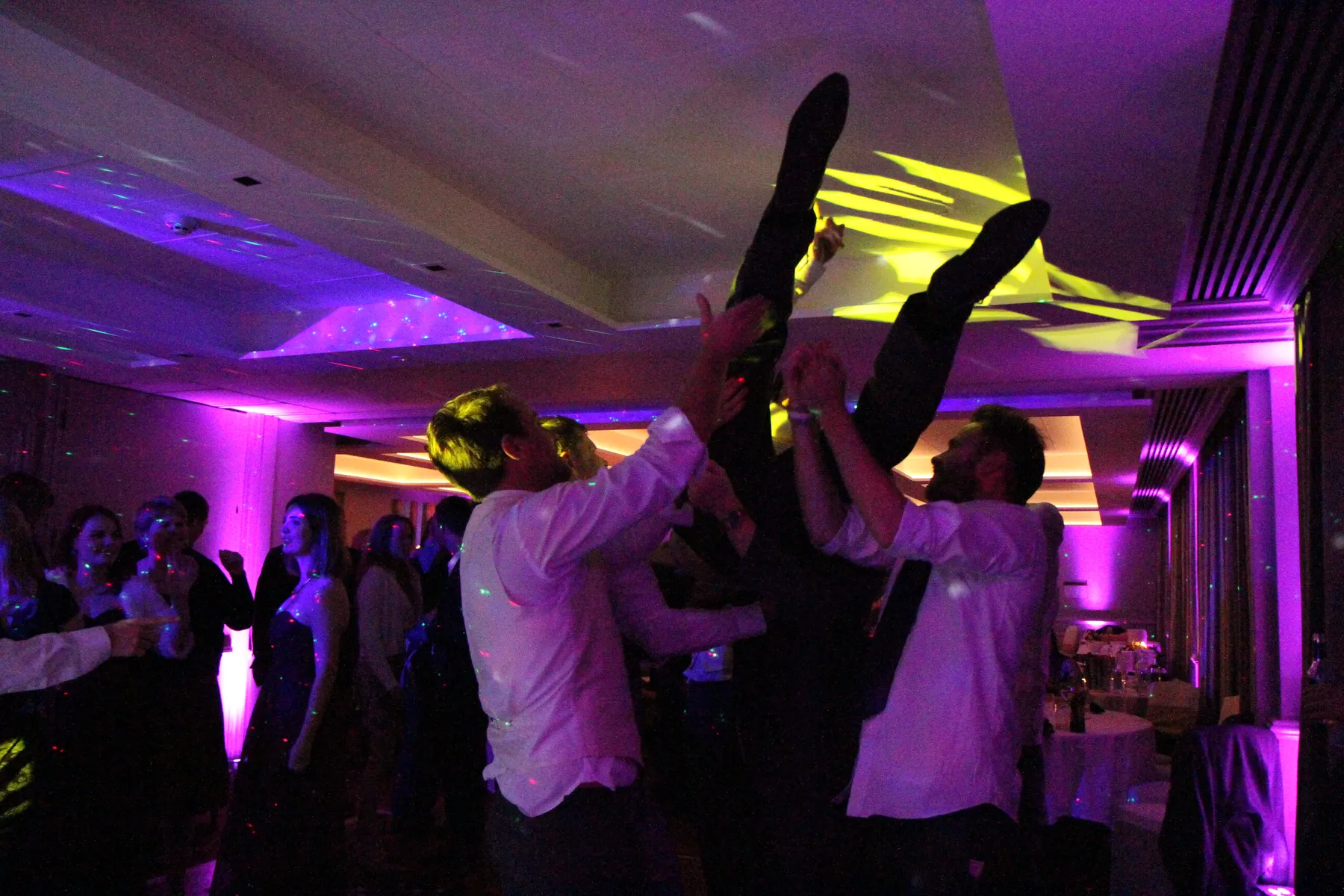 A group of people at a party lift someone upside down in a lively dance floor scene, surrounded by colourful purple and yellow lights, while a wedding DJ Hertfordshire keeps guests in formal attire celebrating in the background.