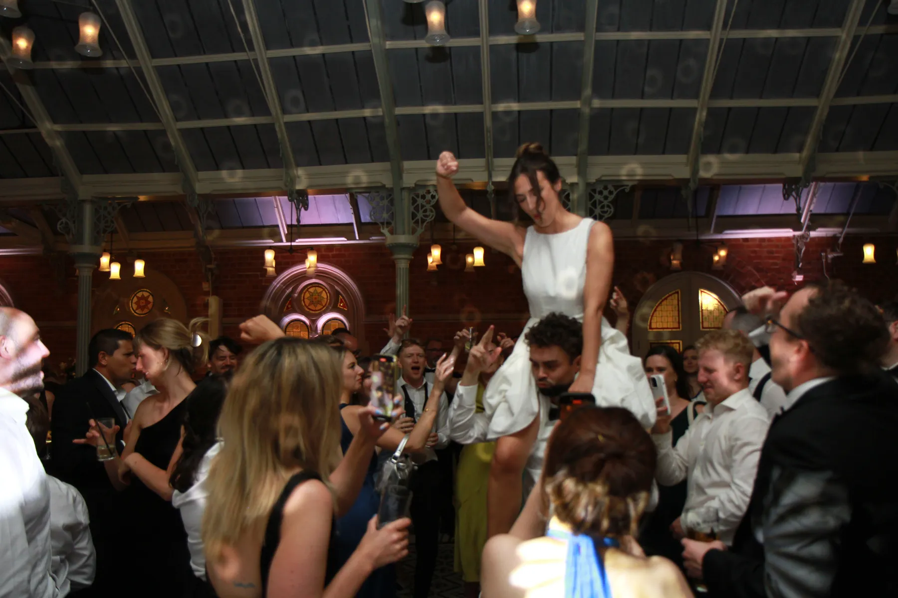 A joyful crowd celebrates at a wedding party at Kilworth House, Leicestershire, with the bride being lifted on the groom's shoulders, raising her fist in excitement under a large glass ceiling with warm lighting.