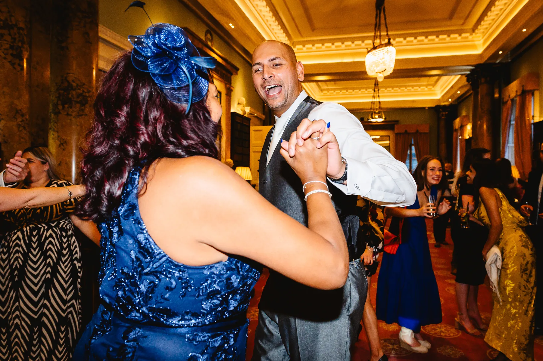 A bridegroom man in a suit and a woman in a blue dress with a matching hat are holding hands and dancing energetically at a lively indoor wedding party at the Athaneum Club, London, with other guests smiling and dancing in the background.